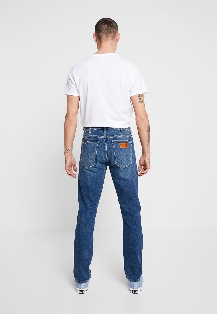 Homme portant un t-shirt blanc et un jean en denim bleu vu de dos, debout devant un fond clair uni.