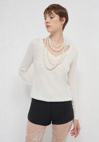 Donna con capelli corti indossa collane di perle a strati, una maglietta bianca a maniche lunghe con scollo a V, pantaloni neri e collant bianchi con motivo di pizzo su uno sfondo neutro.