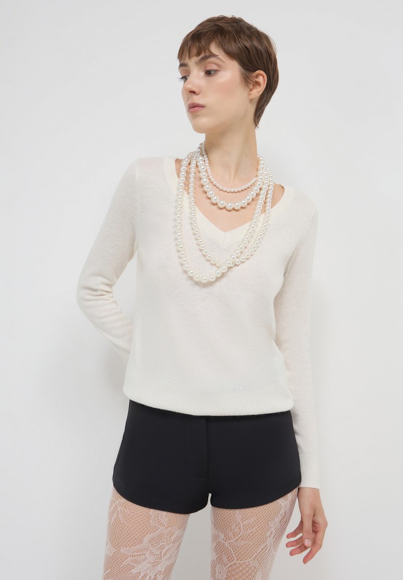 Donna con capelli corti indossa collane di perle a strati, una maglietta bianca a maniche lunghe con scollo a V, pantaloni neri e collant bianchi con motivo di pizzo su uno sfondo neutro.