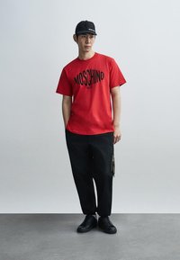 Κόκκινο βαμβακερό μπλουζάκι με το λογότυπο "MOSCHINO" σε μαύρο, σε συνδυασμό με άνετο μαύρο παντελόνι με φερμουάρ στην τσέπη και μαύρα αθλητικά παπούτσια.