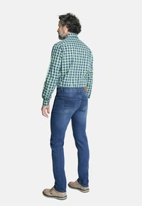 Vista posterior de un hombre que lleva una camisa de cuadros verde y azul, jeans azul oscuro, zapatos marrón claro y un detalle de cuero en la cinturilla.