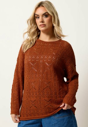 Blonde Frau trägt einen rostfarbenen, gehäkelten Pullover mit geometrischen Mustern und blaue Jeans, hält den Pullover Saum.