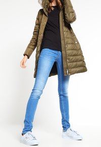 Olivgrön puffjacka med huva och fuskpäls, kombinerad med blå skinny jeans och vita sneakers. Åtsittande design med en glänsande yta.