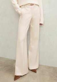 Krémové rozšírené nohavice s hladkou textúrou, s priliehavým pásom a bočnými vreckami, doplnené zlatou sponou na opasku a hnedými podpätkami.