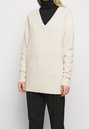 Personne portant un pull ample crème à col en V par-dessus un col roulé noir et un pantalon noir, debout devant un fond clair uni.