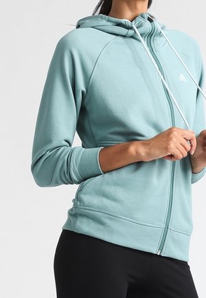 Kvinde justerer snørebåndene på en lyseblå hættetrøje med lynlås og hætte, iført sorte bukser mod en ensfarvet baggrund.