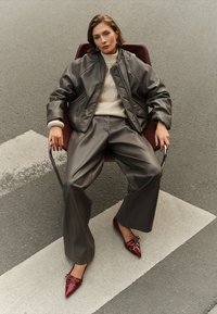 Donkergrijze leren jas en broek gecombineerd met een crème ribgebreide trui, zittend op een stoel, met glanzende bordeauxrode puntige schoenen.