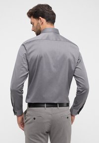 Langärmliges graues Hemd aus glattem Stoff, mit Kragen und geknöpften Manschetten, getragen mit hellgrauen Hosen und einem schwarzen Gürtel.