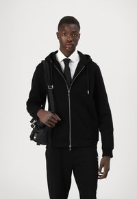 Sweatshirt preto com fecho, de textura suave, emparelhado com uma camisa branca e uma gravata preta. O modelo carrega uma bolsa transversal preta.