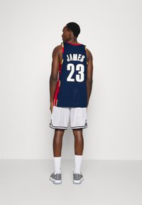 Maglia da basket con base blu navy, dettagli rossi e "JAMES 23" sul retro. Abbinata a pantaloni corti bianchi a righe e scarpe da ginnastica grigie.