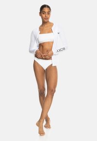 White long-sleeve rash guard with “ROXY” logo on the sleeve, paired with a textured white bikini. Smooth fabric, fitted design.
