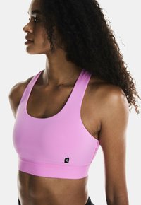 Sujetador deportivo de color rosa claro con una textura suave, tirantes anchos y un diseño ajustado. Presenta una pequeña insignia negra en la parte frontal.