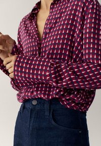 Personne portant une chemise à manches longues à motifs rouges et navy, rentrée dans un jean bleu foncé taille haute, ajustant le poignet de la manche.