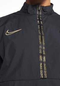 Nike halv-zip jakke i mørk marineblå stof. Har guld accenter, logo og brand-tekst i en kontraststribe langs lynlåsen.