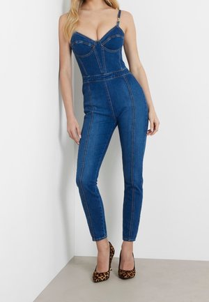 Kvinde iført en tætsiddende blå denim jumpsuit med tynde stropper og leopardmønstrede højhælede sko, der står op ad en hvid væg.
