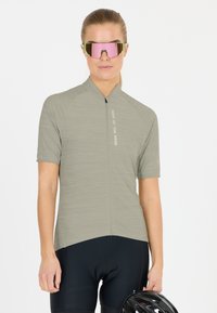 Nainen, jolla on harmaa pyöräilypaita ja mustat shortsit, päässään vaaleanpunasävyiset urheilulasit, pitelee mustaa pyöräilykypärää valkoista taustaa vasten.