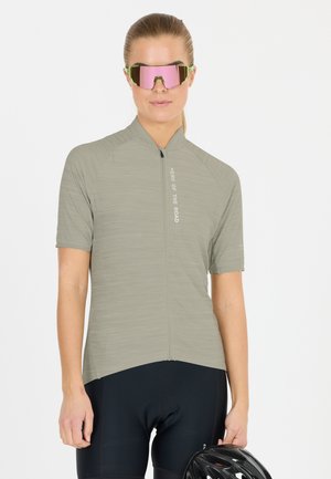 Vrouw in een grijs wielershirt en zwarte korte broek met sportzonnebril met roze tint, die een zwarte fietshelm vasthoudt tegen een witte achtergrond.