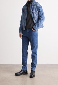 Denim jacket in blue with buttoned front and chest pockets, paired with dark turtleneck and blue jeans. Black ankle boots complete the look.