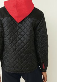 Chaqueta negra acolchada con capucha roja, que presenta una superficie texturizada, costuras diagonales y ventilaciones laterales con cremallera para mayor funcionalidad.