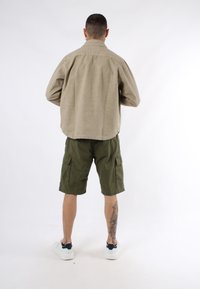 Camicia di cotone beige, pantaloni cargo oliva, sneakers bianche. La vista posteriore mostra le maniche corte, le tasche e una texture sottile. Tatuaggio visibile sulla gamba.