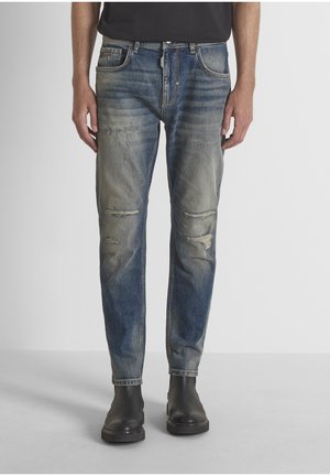 Hombre con jeans azules desgastados y botas negras de cuero hasta el tobillo, de pie frente a un fondo claro y liso.