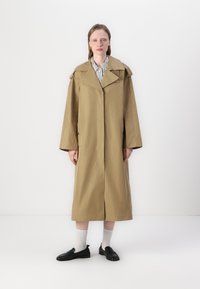 Beige trenchcoat med brede ærmer og en stor krave, med knapper og slids bagpå, båret over en stribet skjorte og parret med sorte sko.