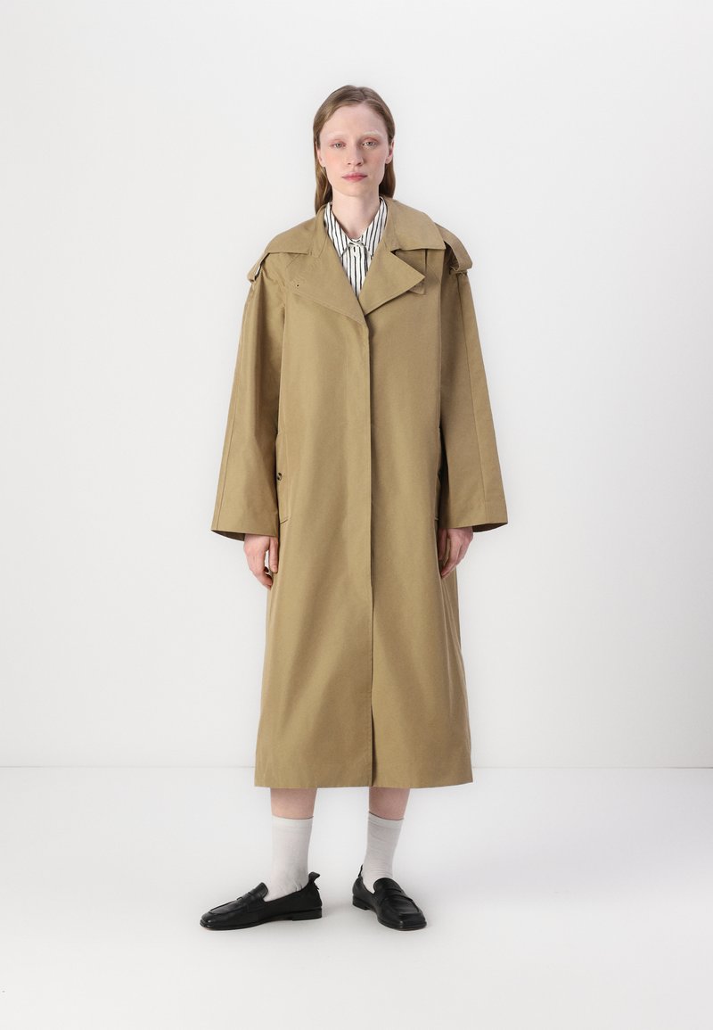Beige trenchcoat med brede ærmer og en stor krave, med knapper og slids bagpå, båret over en stribet skjorte og parret med sorte sko.