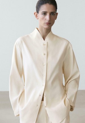 Femme portant une chemise ample crème à manches longues boutonnée et un pantalon assorti, debout les mains dans les poches devant un fond neutre.