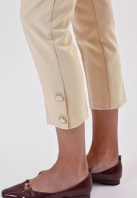 Pantaloni beige a cropped con bottoni laterali e una texture liscia, abbinati a scarpe lucide bordeaux con cinturino e decorazione rotonda.