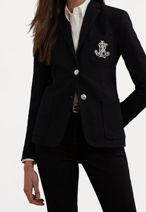 Mujer con blazer negro entallado con botones plateados y monograma bordado en el bolsillo del pecho, combinado con camisa blanca y pantalones negros.