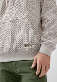 Jasnoszara, teksturowana bluza z kapturem z tkanym wzorem, kieszenią typu kangur oraz ściągaczami na mankietach. Posiada naszywkę z logo z napisem "Elevated Casual Est. 1969."