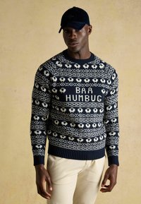 Marineblauer Strickpullover mit weißen Schafen und geometrischen Mustern, versehen mit dem auffälligen Text "BAA HUMBUG". Gerippte Bündchen und Saum.