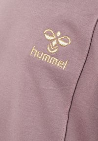 Logo brodé en or représentant une abeille avec le texte "hummel" sur un tissu mauve, mettant en valeur la texture et le détail de la couture du textile.