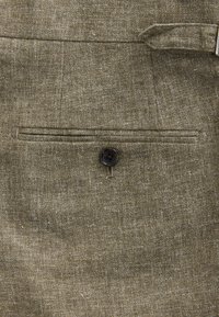 Olivengrønt stof med en tekstureret vævning. Indeholder en vandret lomme med enkelt sort knaplukning og synlige syningsdetaljer.
