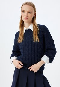 Maglione lavorato a maglia blu navy con texture a treccia, abbinato a una camicia bianca con colletto. Vestibilità rilassata e maniche oversize.