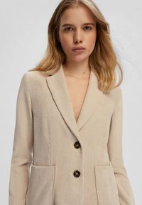 Blazer in corduroy beige con revers a punta, due tasche frontali e tre bottoni marroni. Texture liscia con motivo a righe verticali.