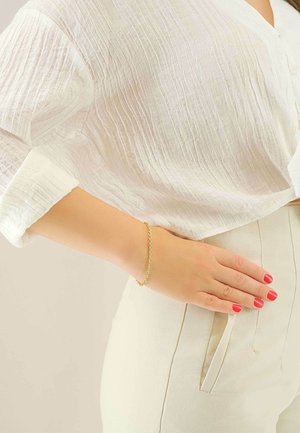 Bracciale in catena d'oro su un polso, abbinato a una blusa bianca a trama e pantaloni alta vita di colore chiaro. Smalto rosso brillante visibile.