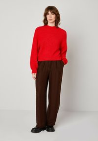 Raudonas megztinis su aukšta apykakle, derinamas su laisvai krentančiomis rudos spalvos raukinėtomis kelnėmis ir juodais stambiais batais. Paprastas ir tekstūruotas dizainas.