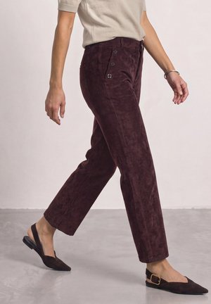 Femme marchant portant un pantalon en velours côtelé violet foncé avec boutons sur le côté, un haut beige à manches courtes et des ballerines slingback pointues foncées avec une boucle.
