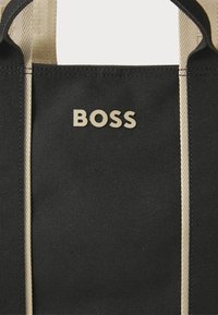 Borsa tote in tela nera con minuteria dorata. Presenta due strisce verticali color beige e la parola "BOSS" in rilievo con lettere dorate.