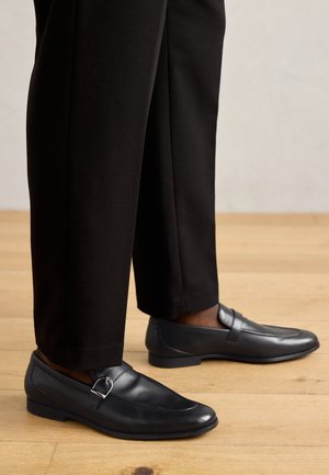 Schwarze Leder-Loafer mit silberner Schnallendetail, getragen zu schwarzen, maßgeschneiderten Hosen auf einem Holzboden.