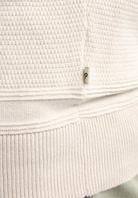 Cream-colored textured fabric with a waffle pattern, featuring a ribbed cuff and a small branded tag at the seam for detail.