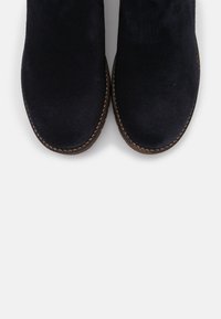 Botines de ante azul marino con la puntera redondeada y costuras marrones a lo largo de la suela de goma, mostrando una textura suave y un diseño minimalista.