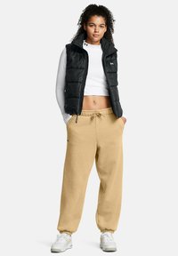 Svart pufferväst över en vit långärmad tröja, kombinerad med beige sweatpants och vita sneakers. Har dragkedjestängning och elastiska ärmslut.