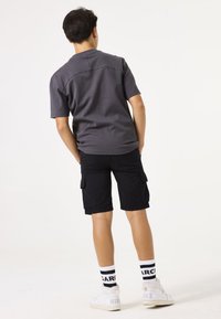Graues kurzärmeliges Hemd, schwarze Cargo-Shorts mit Seitentaschen, weiße Sneaker mit Logo und gestreifte schwarz-weiße Socken.