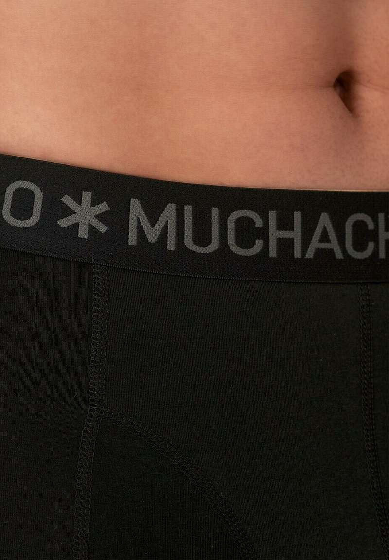 Boxer breves de algodón negro con una amplia cinturilla negra que presenta la inscripción "MUCHACHO" en gris. La tela parece suave con costuras planas.