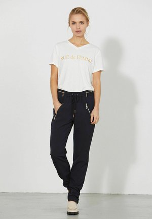 Blondínka stojí pri bielej stene, oblečená v bielom tričku "RUE de FEMME", čiernych nohaviciach so šnúrkou a zlatými zipsami a béžových mokasínach.