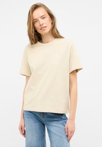 Beige cotton t-shirt with short sleeves, round neck, and a small shell graphic on the left chest. Worn with light blue jeans.