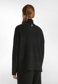 Schwarzer oversized Pullover mit hohe Kragen, weichem Material, geripptem Saum und einem kleinen rosa Logo auf der Rückseite nahe dem Kragen.