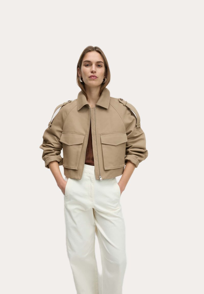 Beige cropped jack met ritssluiting, gevoerde schouders en twee voorke pockets. Draagt over een bruine top en gecombineerd met witte broek.